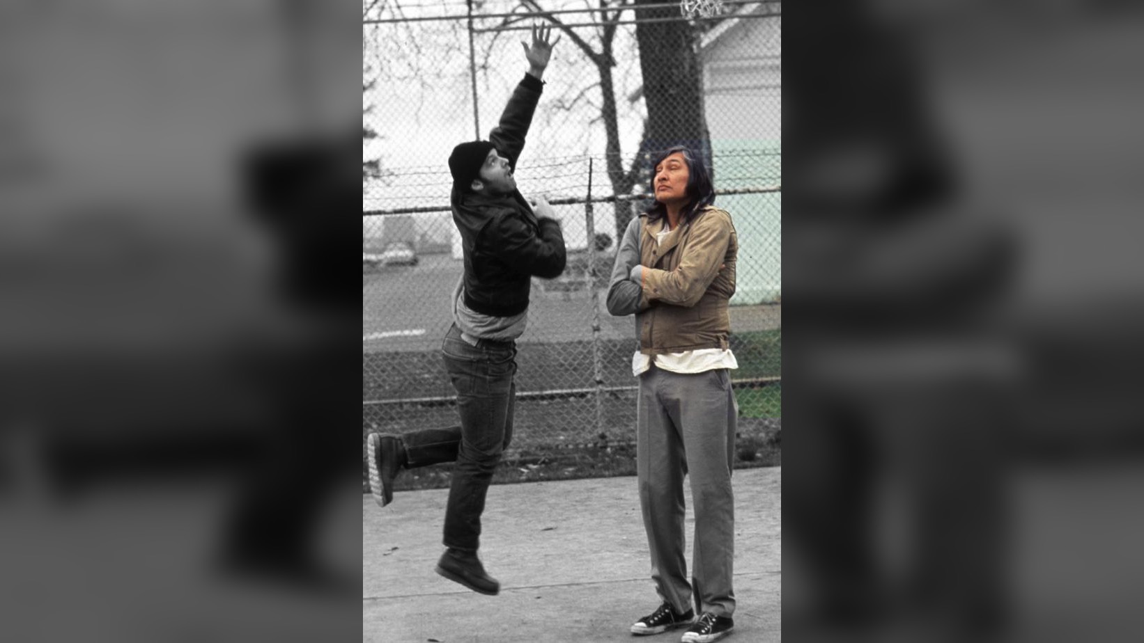 Image from the 1975 film One Flew Over the Cuckoo's Nest. Murphy, played by Jack Nicholson, throws a basketball into a hoop. Chief Bromden, played by Muscogee Nation actor Will Samson, watches in silence, his arms crossed.